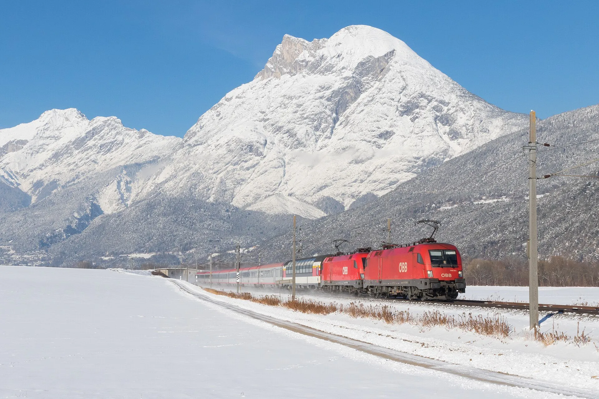 A red ÖBB railjet locomotive hauling the EC Transalpin across a snowy valley floor below a snow-covered peak.