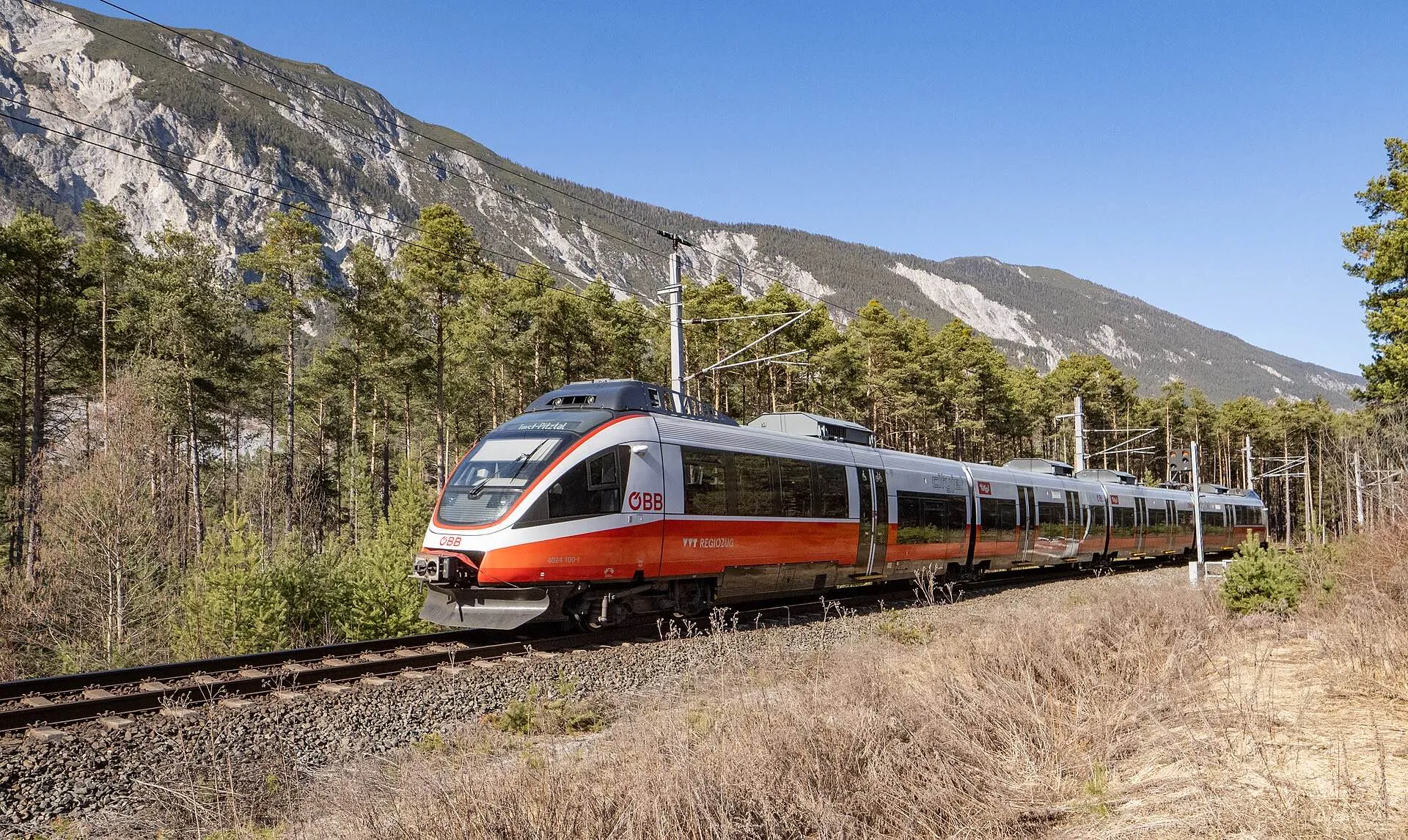 A red and white ÖBB Talent regional train running through pine forest between Ötztal and Roppen.