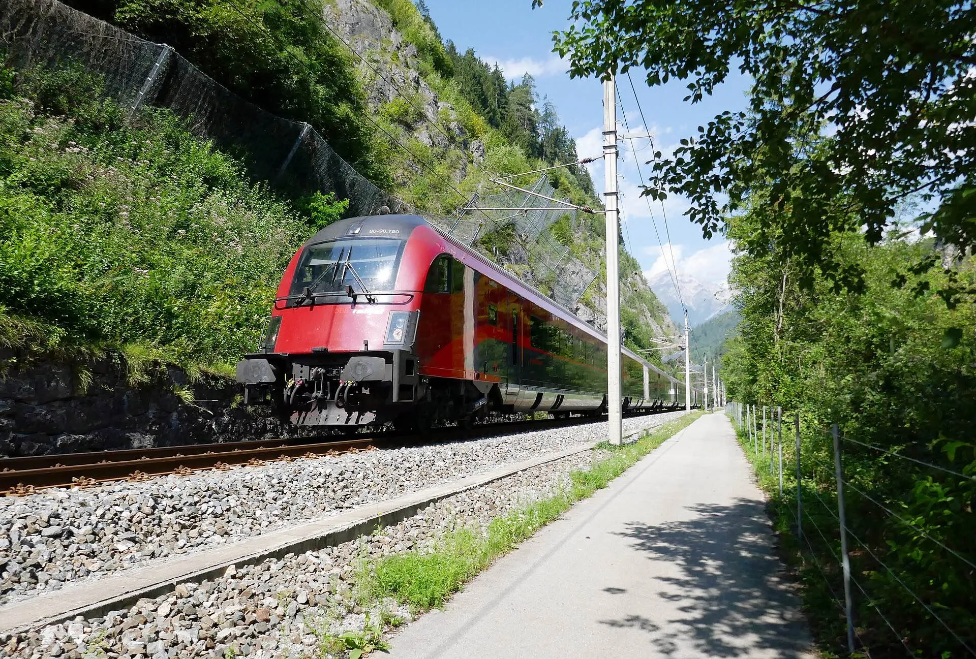A red railjet passing a wall of rock and a footpath on the Arlbergbahn in summer.