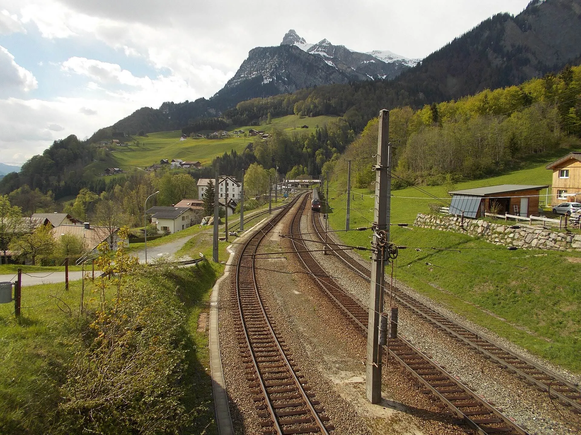 The twin tracks of the Arlbergbahn curving through green meadows at a village station below the Roggelskopf.