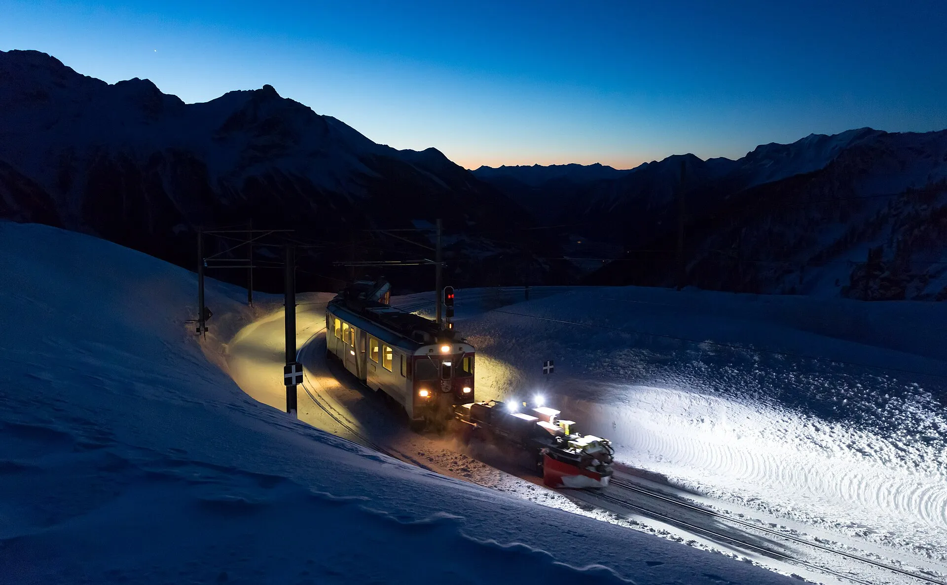 A lit Bernina line railcar with a snow plough working at night in deep snow near Alp Grüm.