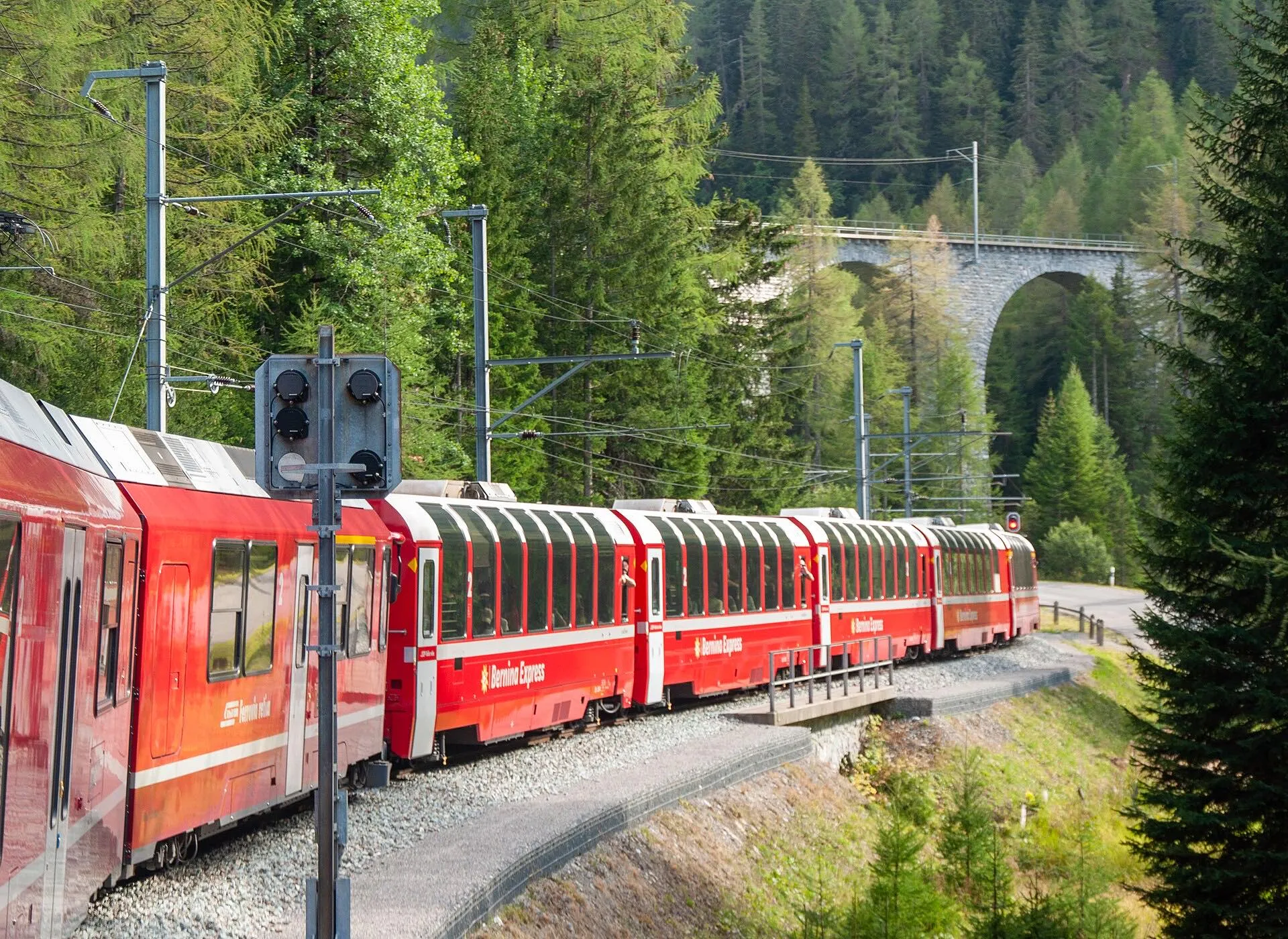 The panoramic Bernina Express train curving past a stone viaduct and larch forest.