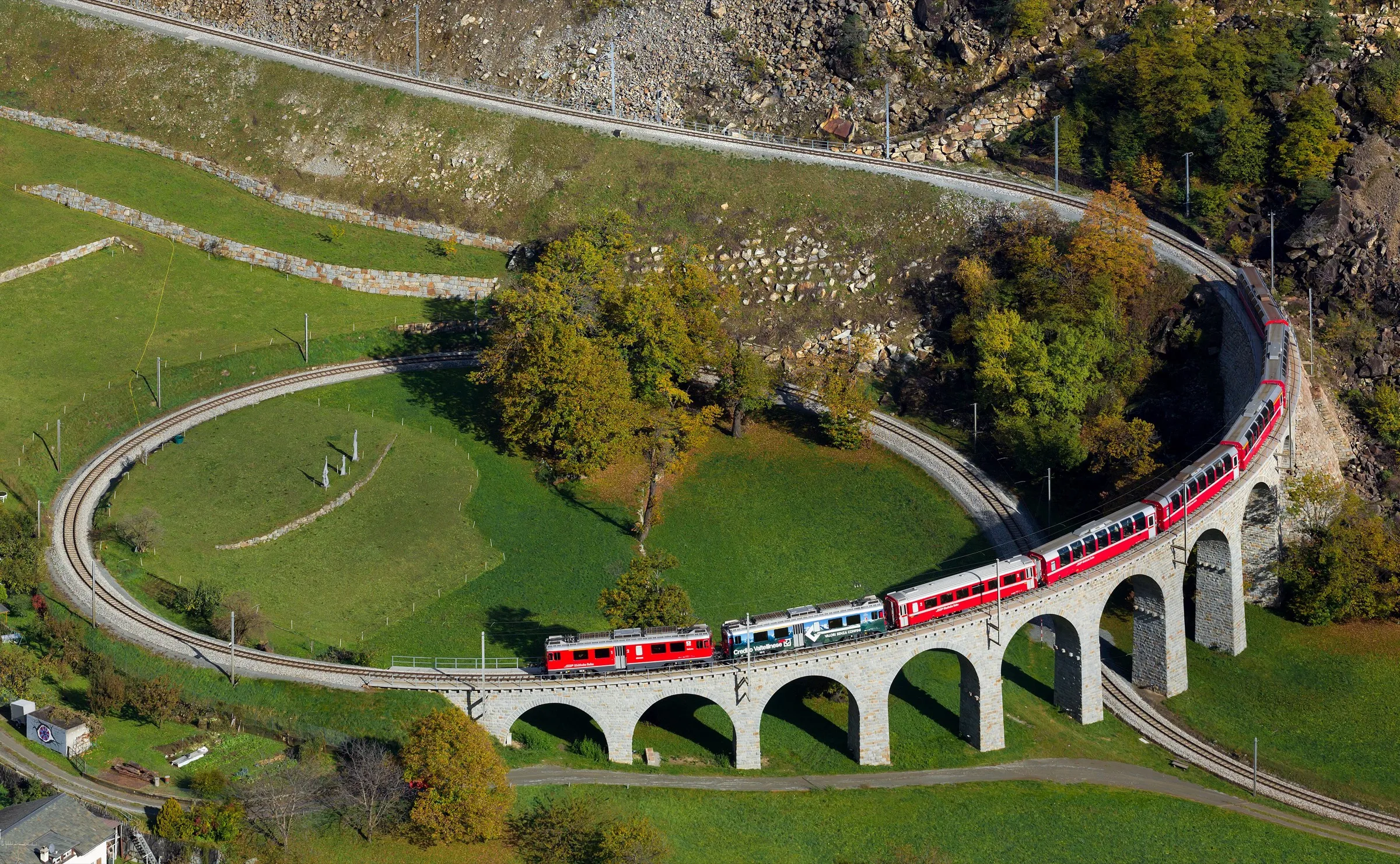 A red Bernina line train curving down the Brusio spiral viaduct between meadows and stone walls.