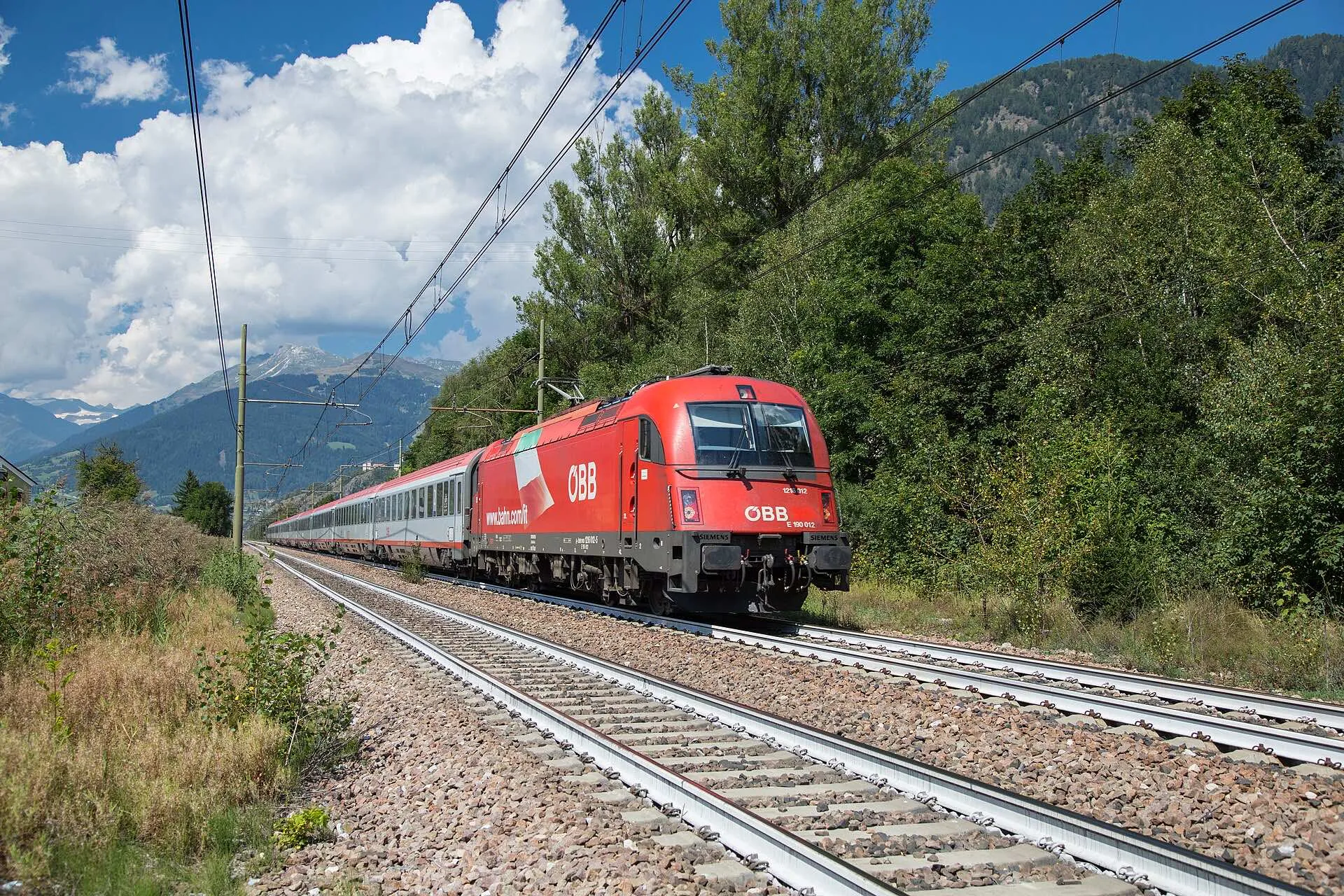 A red ÖBB Eurocity train running past pine trees near Campo di Trens with peaks behind.
