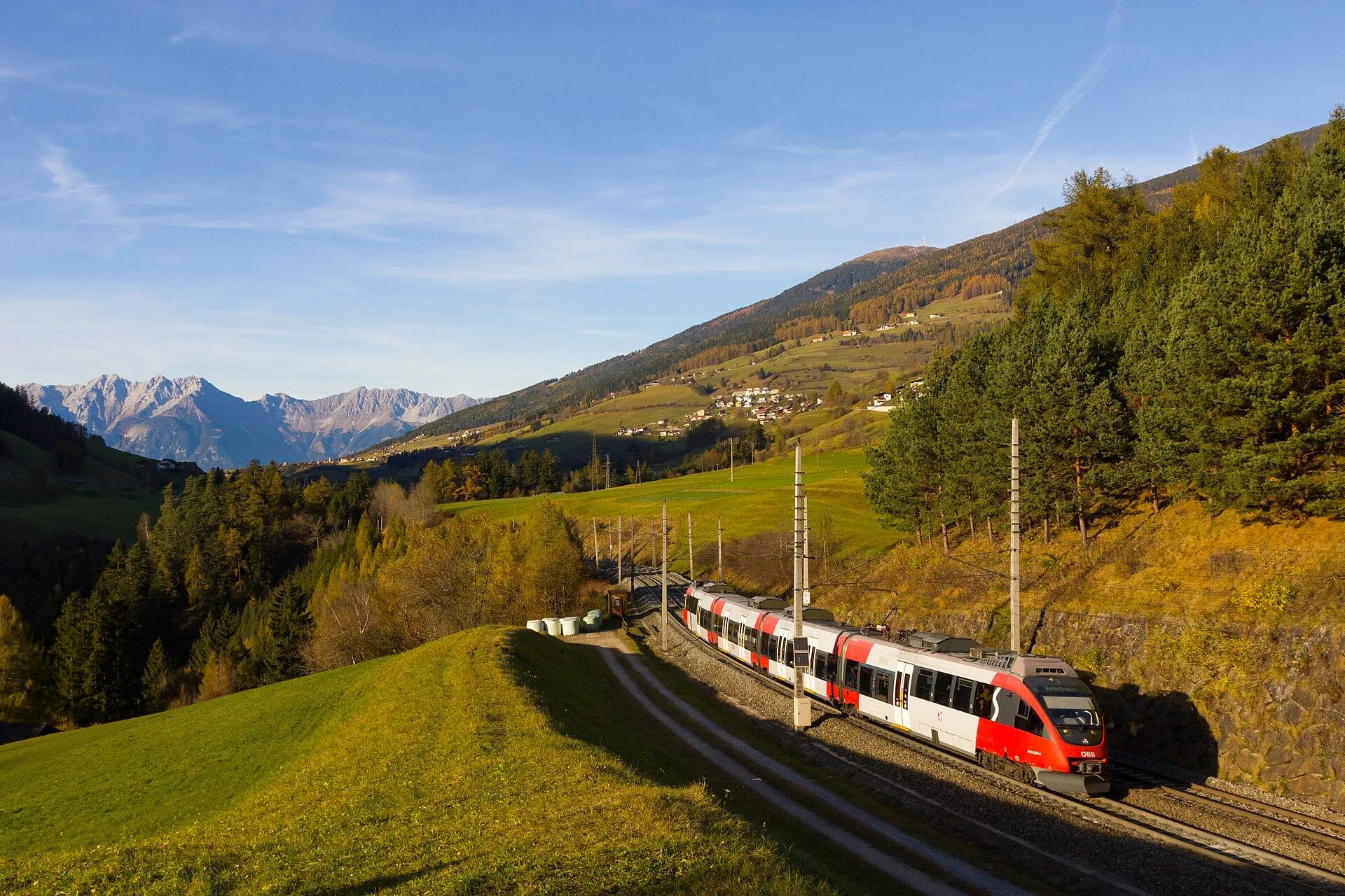 A red and white ÖBB Talent train rounding a curve through autumn slopes near Matrei.