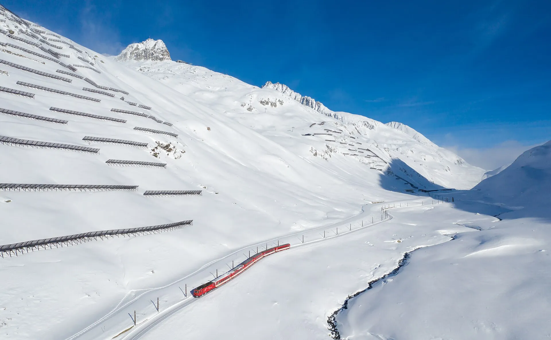 A Matterhorn Gotthard Bahn train climbing through deep snow towards the Oberalp Pass under a blue sky.