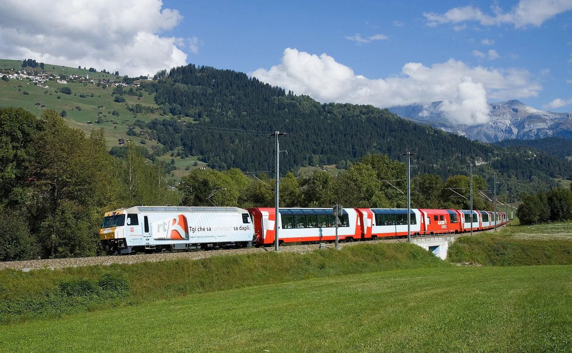 A white Rhaetian Railway locomotive hauling the Glacier Express past forests near Ilanz.