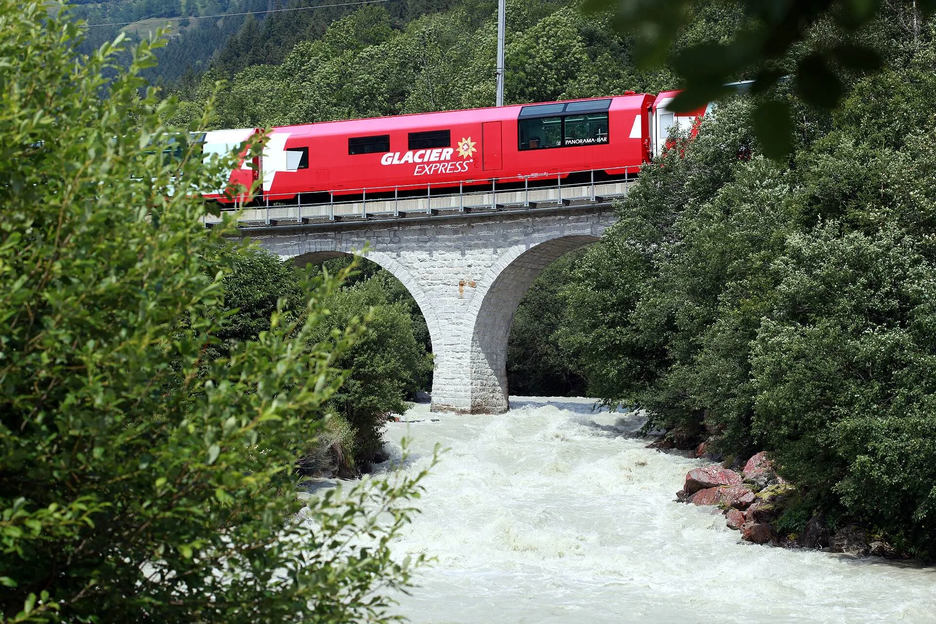 The Glacier Express crossing an arched stone bridge over a rushing mountain river.