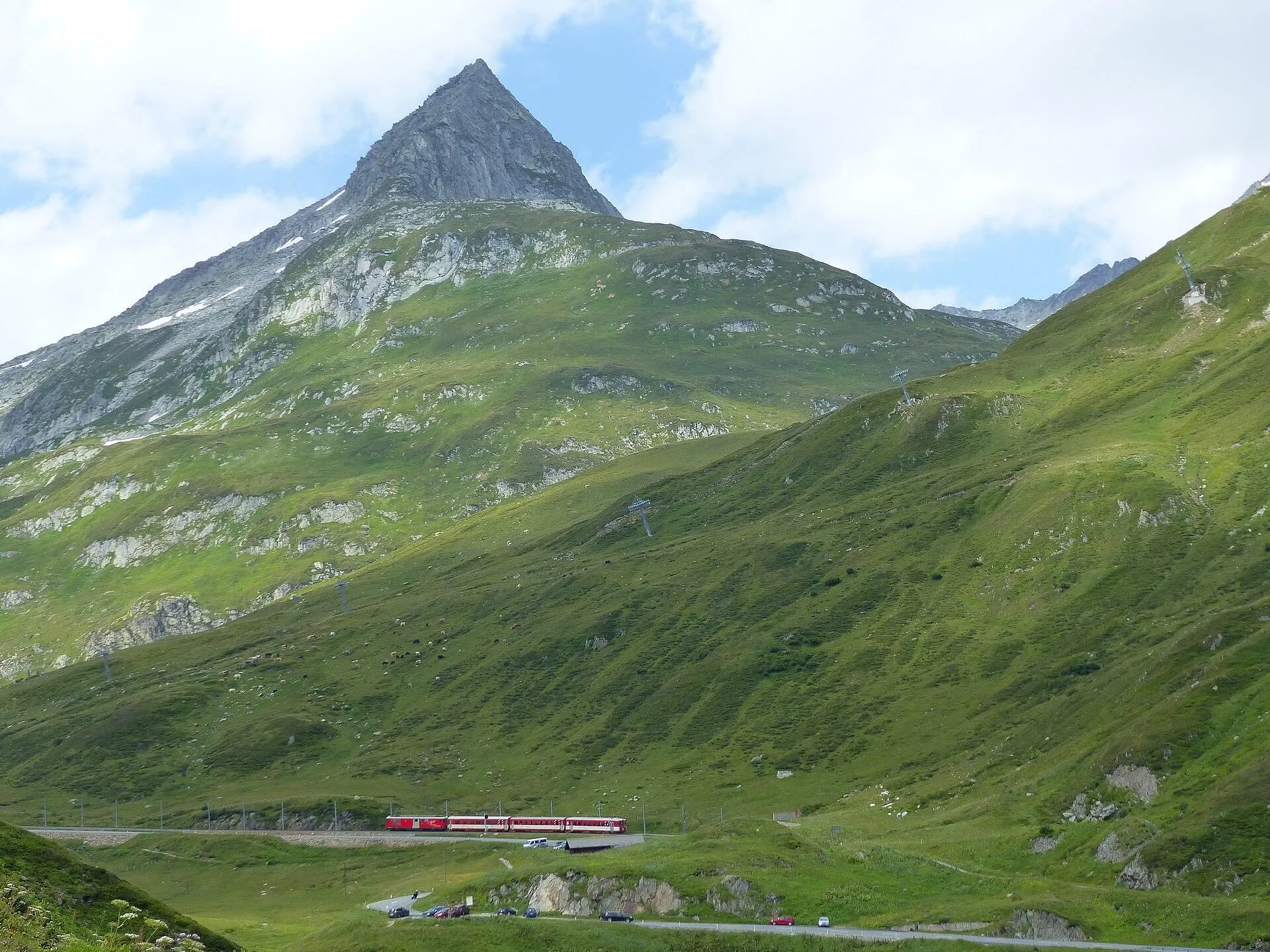 A small red train crossing the green valley below the pointed Piz Tiarms near the Oberalp Pass.