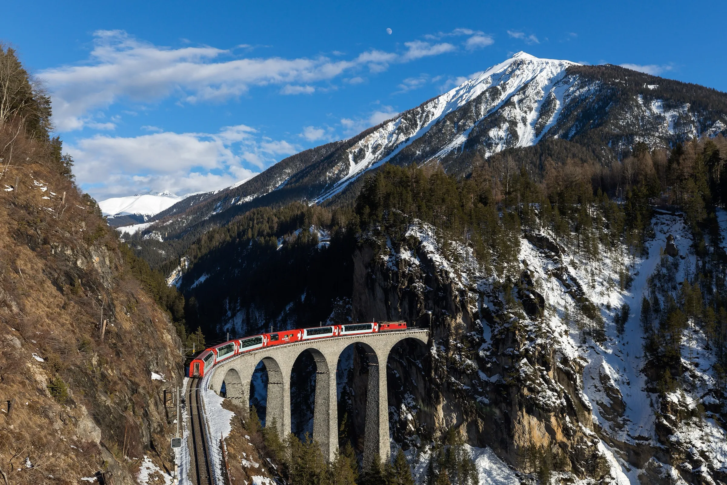 The red Glacier Express crossing the Landwasser viaduct in winter with snow-covered mountains behind.