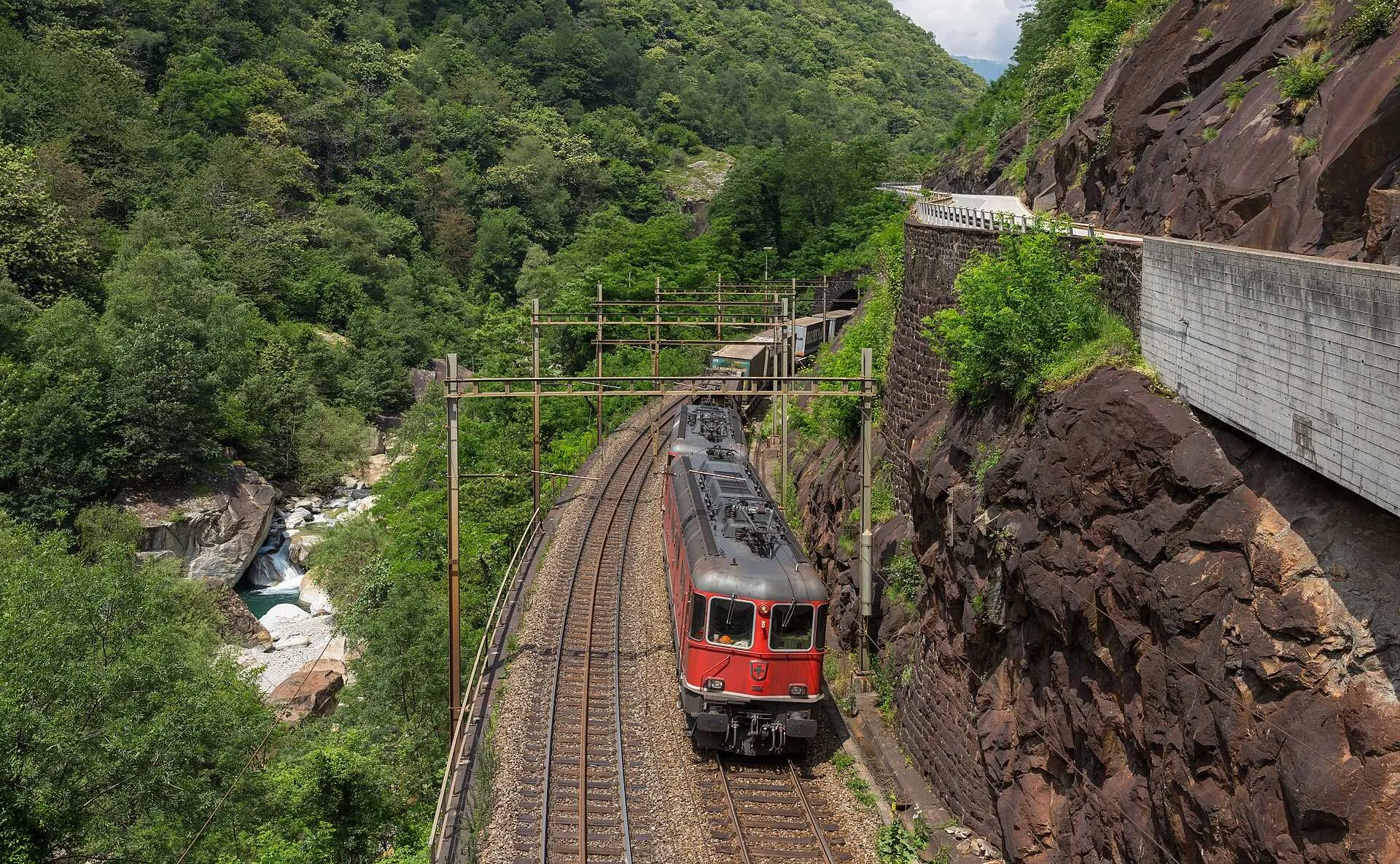 A red SBB locomotive climbing along a rock wall above the river in the Biaschina gorge.