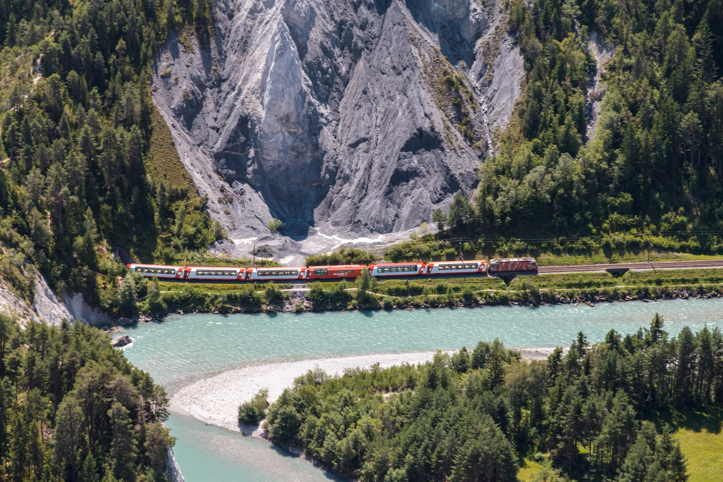 The Glacier Express running along the turquoise Rhine through the Rhine gorge below pale limestone cliffs.