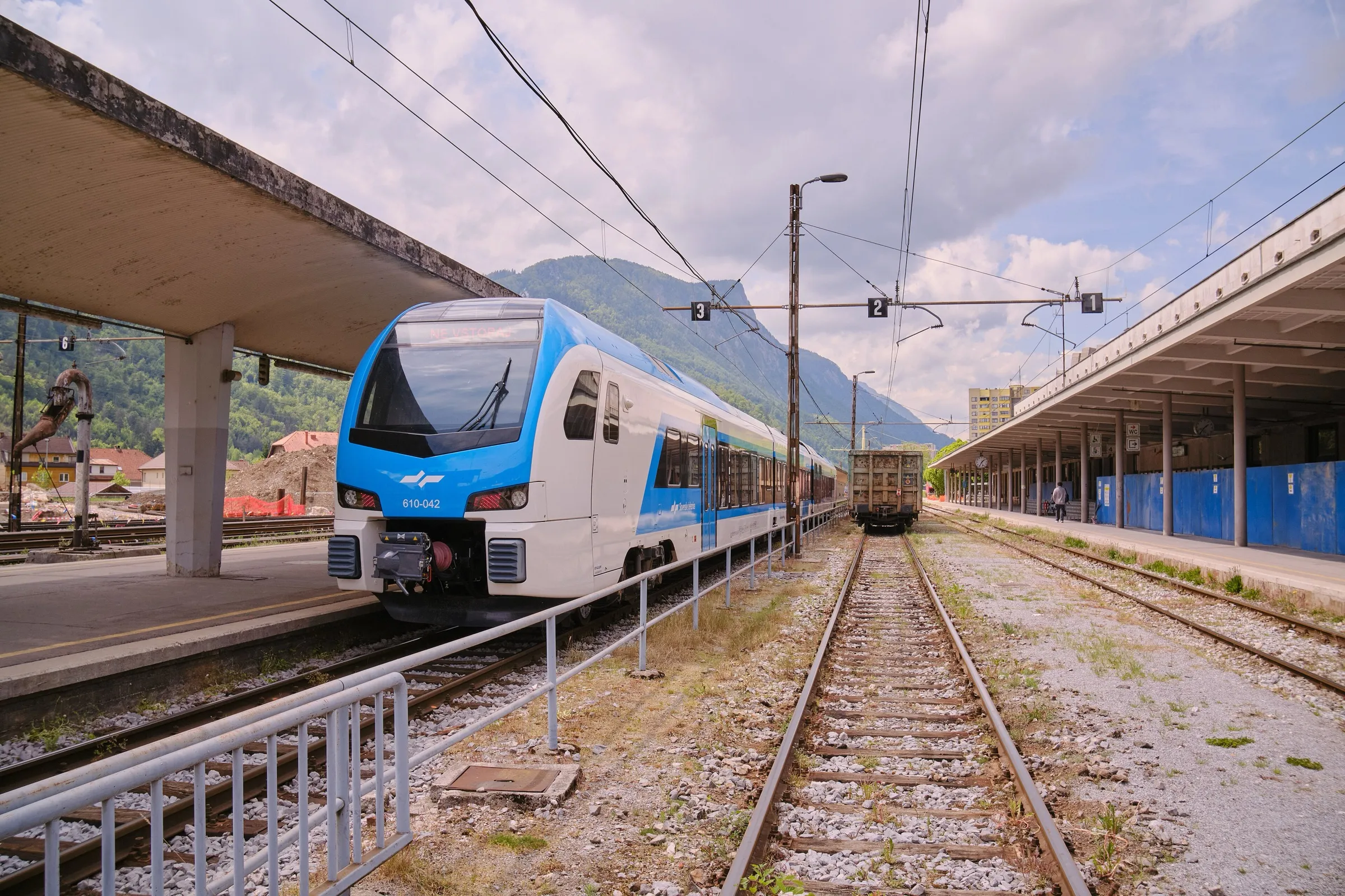 A blue and white Stadler Flirt train at the platform of Jesenice station with green hills behind.