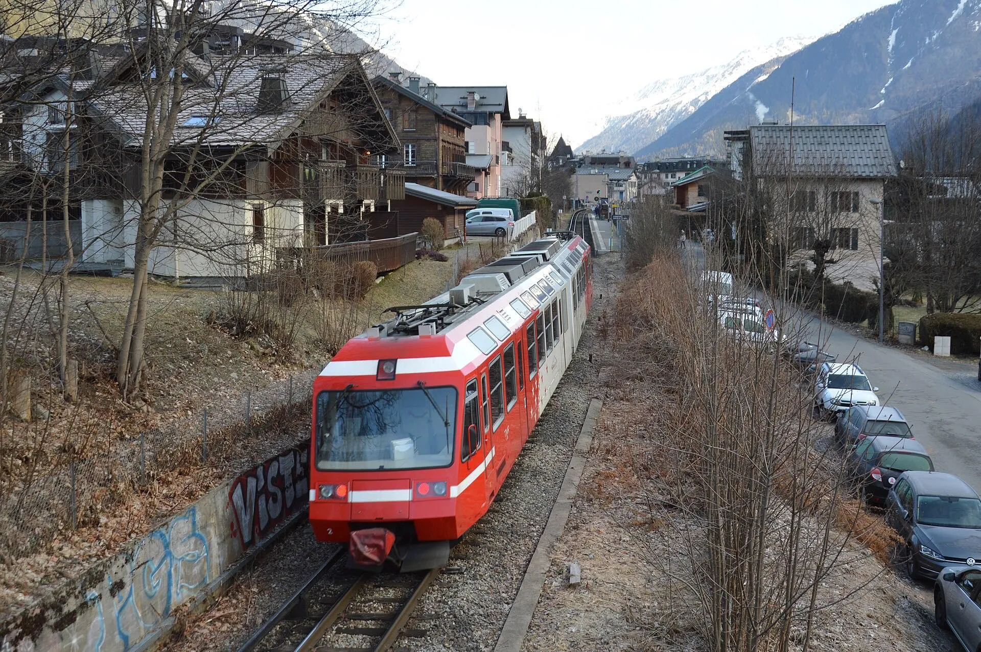 A red TER train curving into Chamonix Aiguille du Midi station between chalets and trees.