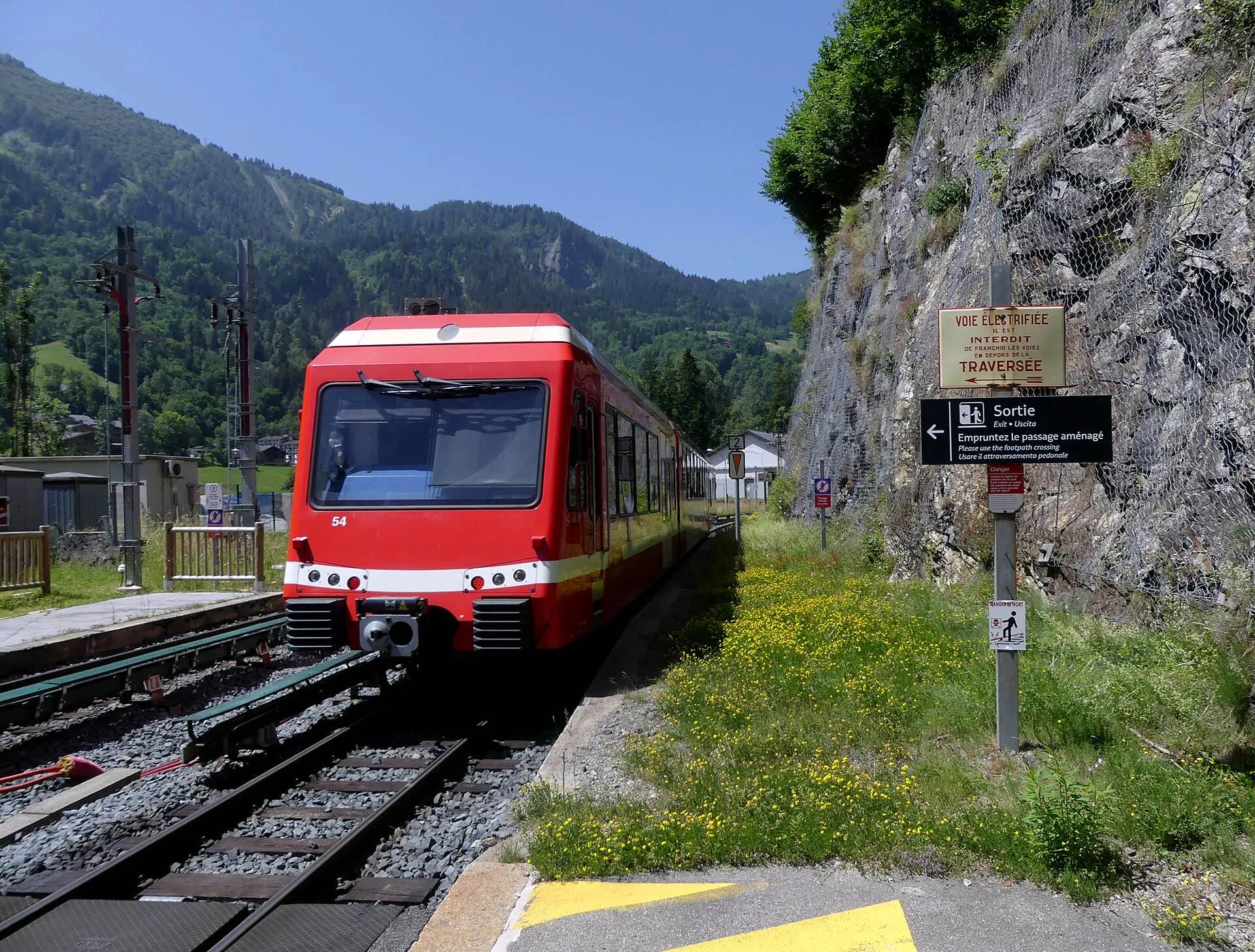 A red TER train leaving Les Houches on a curve lined with trees and chalets.
