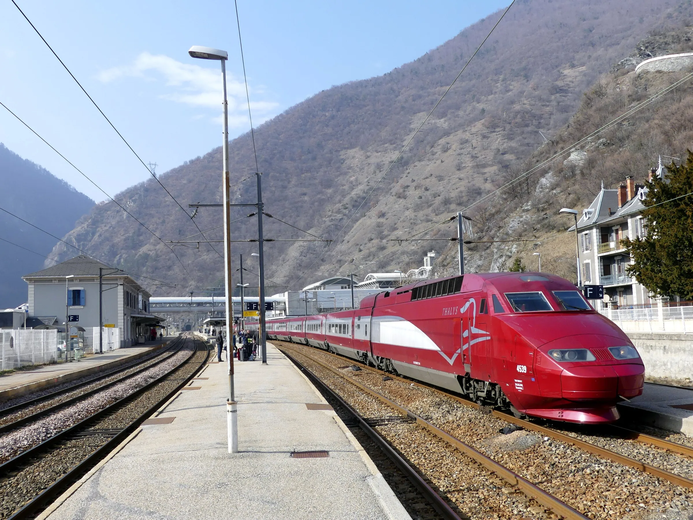 A red Thalys high-speed train stopped at Moutiers station in winter with steep wooded slopes behind.